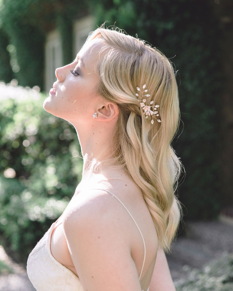 A model raises her face to the sun; her blonde hair is styled in soft waves and a hair pin styled above the ear. The hair pin is gold, with blush flowers and crystals. She is wearing stud earrings with a cluster of crystals above a blush pearl.