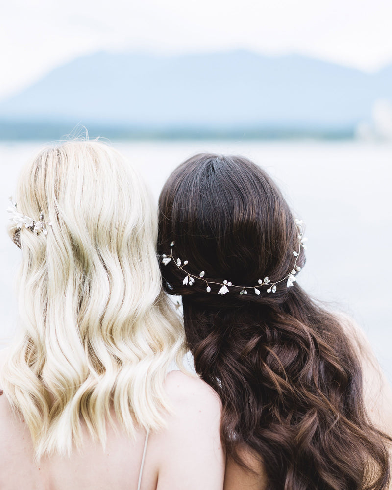 Two models face the ocean with their backs to the camera. The dark-haired model on the right wears the Belle Fleur Hair Vine styled across the back of her half-up hairstyle. The hair vine has flowers, pearls, and crystals.