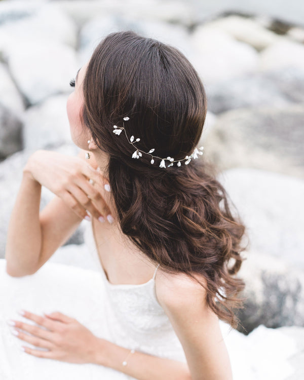 A bride poses on the rocky shore. She is wearing a hair vine styled in the back of her half-up hairstyle. The hair vine has flowers, pearls, and crystals.
