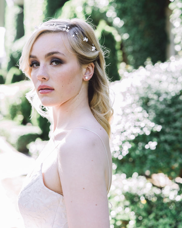 A blonde model with smoky eyes gazes directly into the camera. She is wearing a delicate hair vine made of flowers, pearls, and crystals. She is also wearing classic pearl studs.