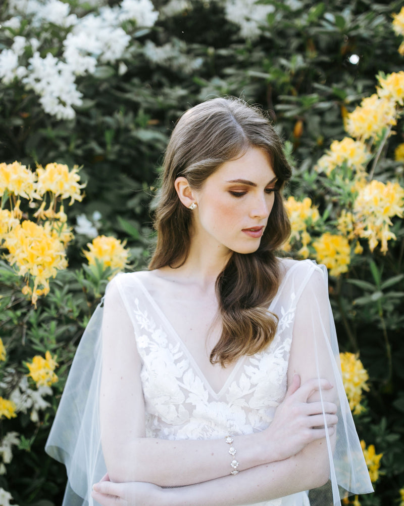 A bride poses in a tulle bridal cape, wearing the Celestial Pearl Cluster Bracelet in gold and matching pearl stud earrings.