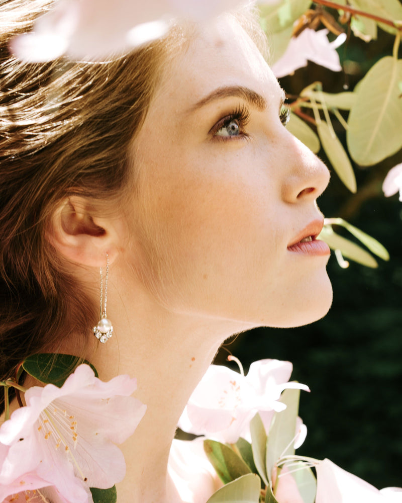 A model poses with white flowers all around her. She is wearing the Celestial Threader Earrings in silver with cream pearls.