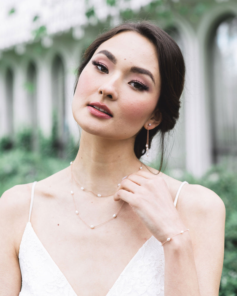 A model is wearing a pearl jewellery set. She is wearing a layered pearl necklace with matching dainty pearl bracelet, along with long drop earrings with a trio of pearls.