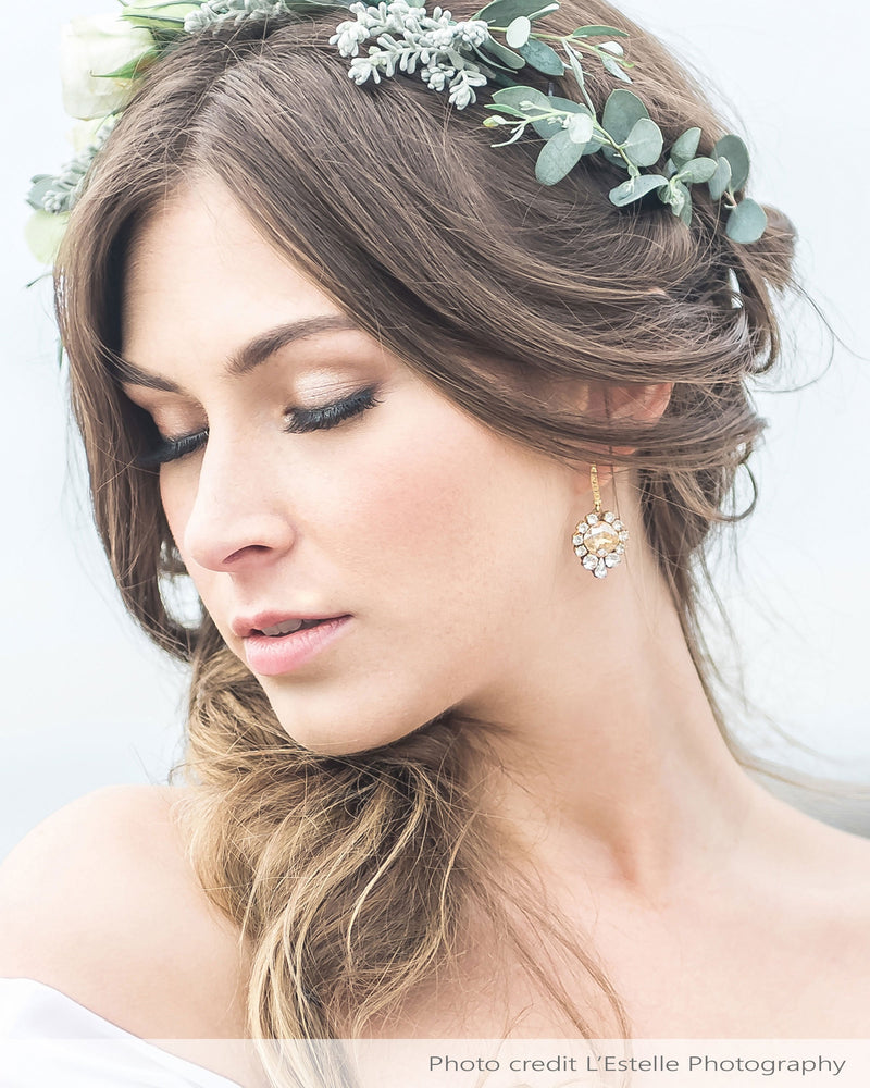 A model with a floral crown wears the Enchanted Crystal Drop Earrings in gold with champagne crystal centres. She is wearing the pave crystal hook version.