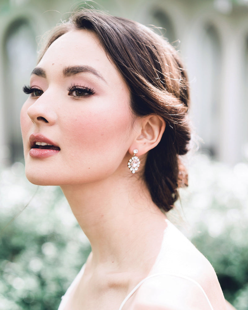 A model smiles as she wears the Enchanted Crystal Drop Earrings in gold with blush crystal centres. Styled with a low bridal updo with twists.