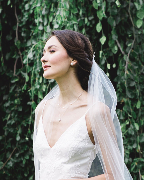 A bride wears a simple veil and models the Halo Pearl Layered Necklace in silver with freshwater pearl and the matching Halo Pearl Stud Earrings.