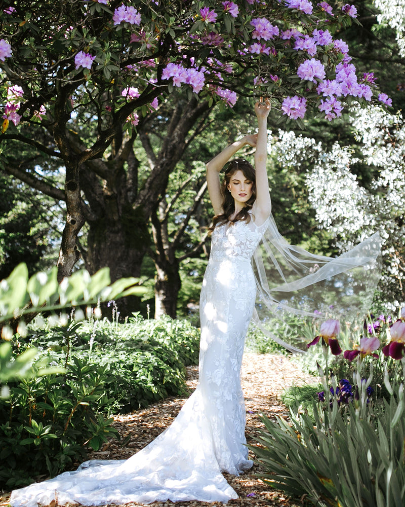 modA bride stands in a garden with blooming shrubs. Her waltz length veil floats in the air behind her.el wearing lily short plain wedding veil