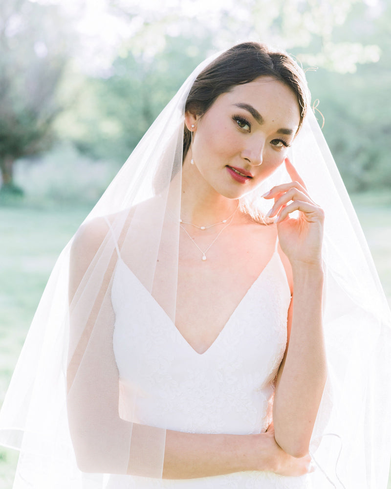 A bride wears a modern pearl jewellery set with Long Pearl Teardrop Earrings and a Layered Pearl Drop Necklace, paired with a two-layer veil with a ribbon trim.