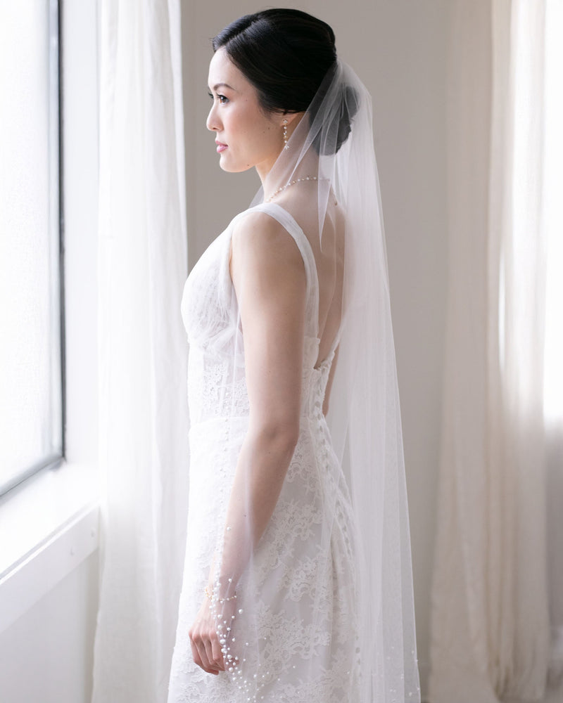 A bride gazes out a window wearing the Kalina Pearl Veil in fingertip length.