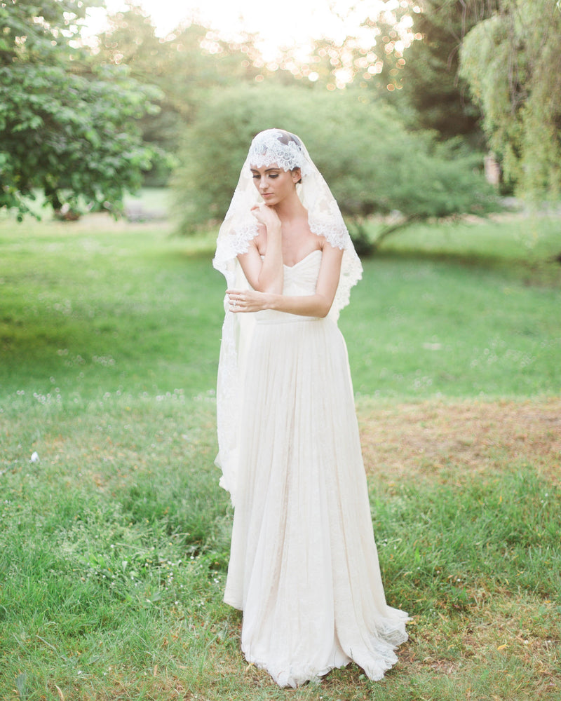 A model stands in a sunlit field, wearing a chantilly lace mantilla veil with wide lace border.
