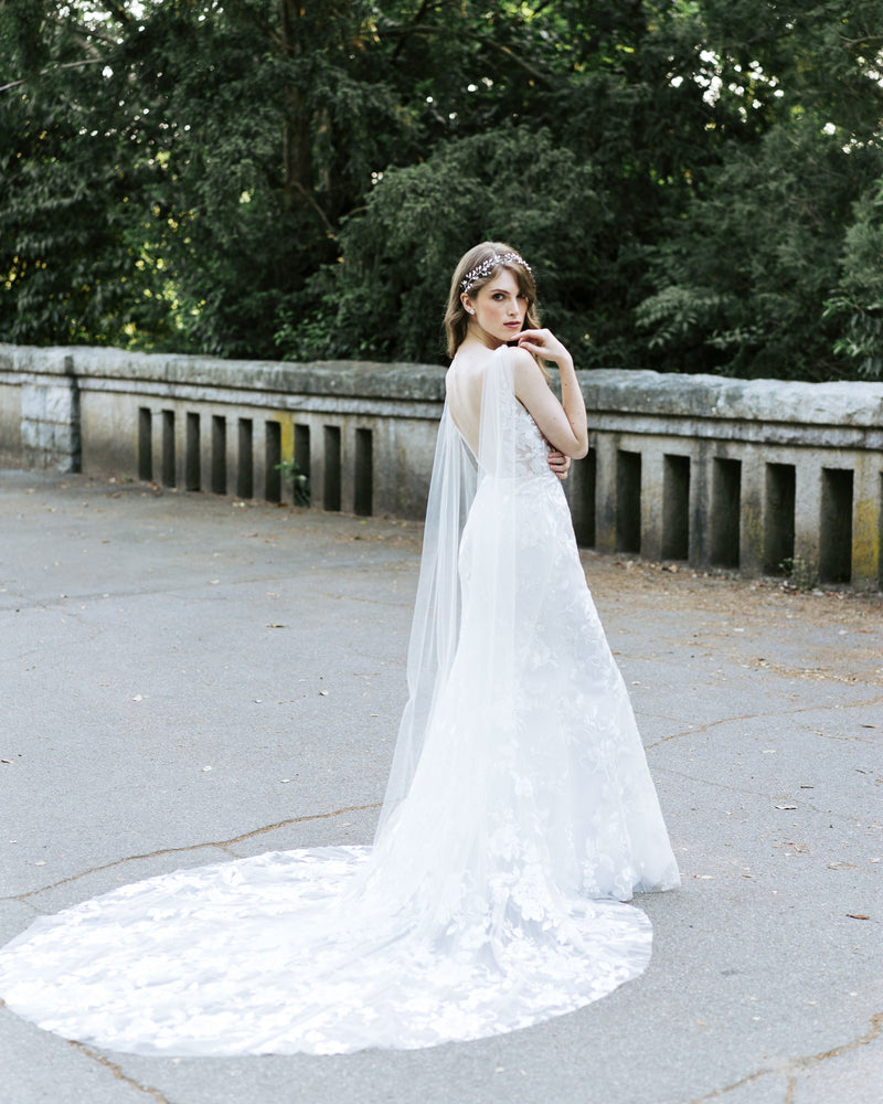 A bride wears the Aster Cape Veil in chapel length. The veil attaches to her V-neck straps in the back.