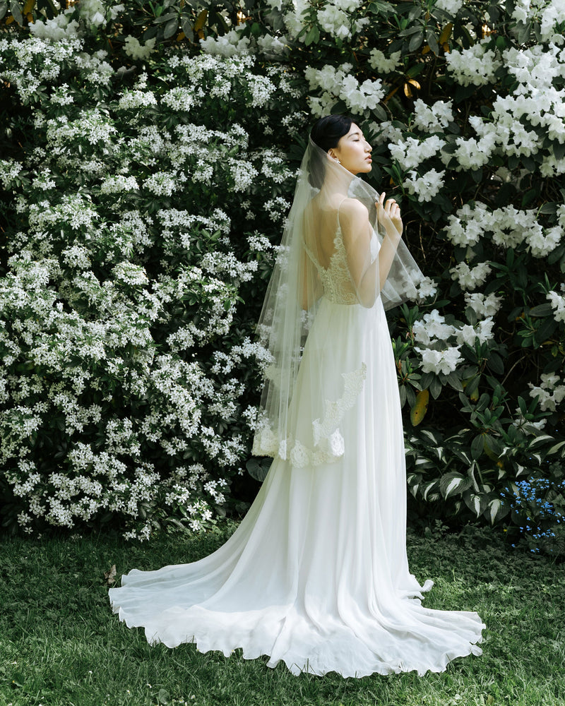 A bride stands in a front of blooming hedges, wearing the Azalea lace bridal veil.