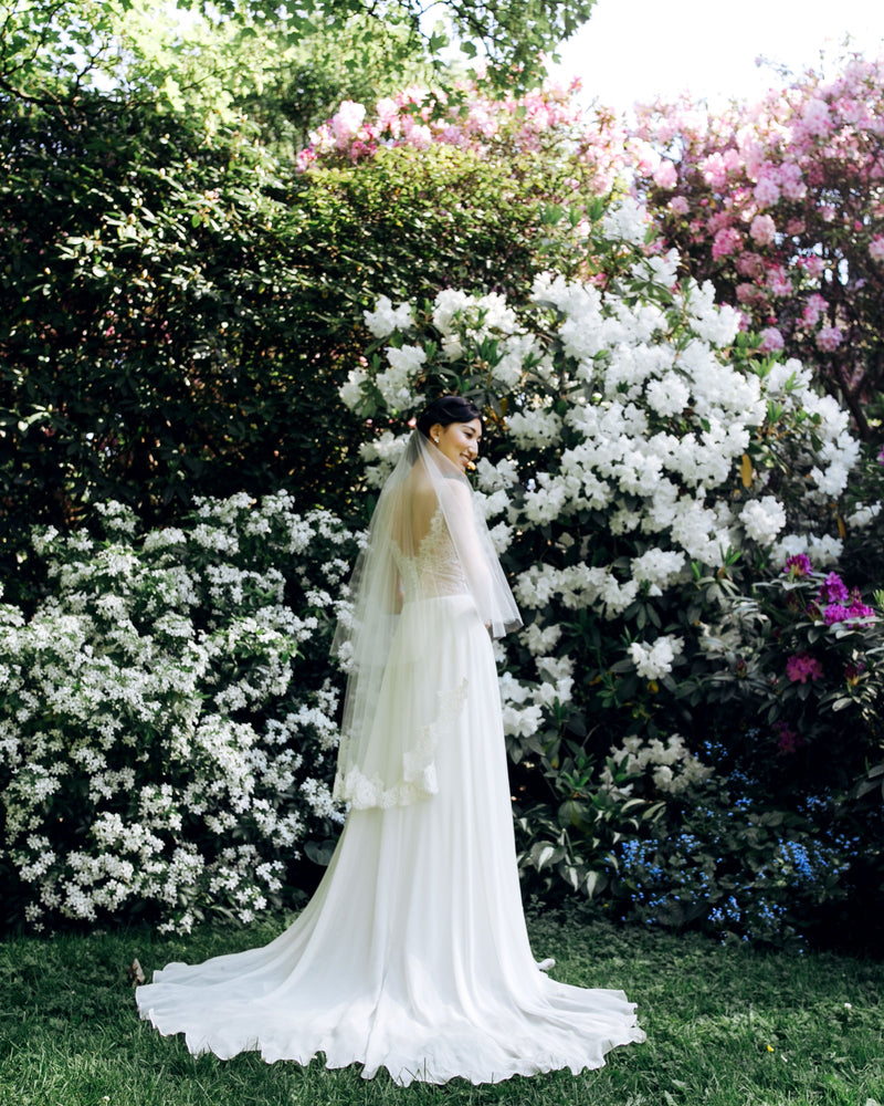 A bride stands in a front of blooming hedges, wearing the Azalea lace bridal veil.