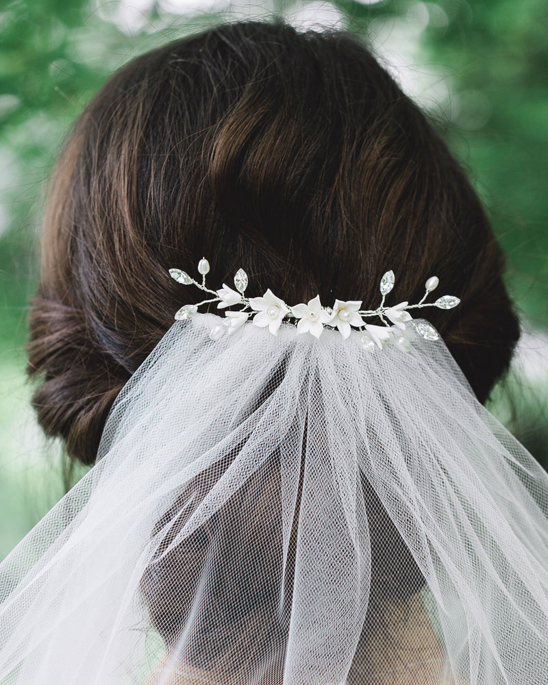 Close-up of a bride with dark hair and low updo wearing a bridal hair comb with crystals, white flowers, and pearls over a veil.