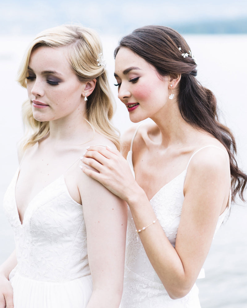 Two models pose together, both wearing bridal accessories with flower details. On the left, the model wears tiny drop earrings with white flowers and pearls. On the right, model wears delicate statement earrings and a dainty pearl bracelet.
