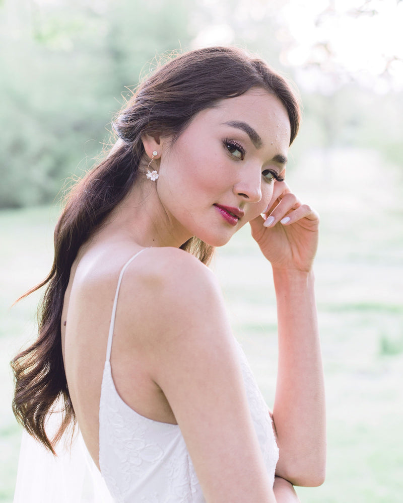 A bride with dark hair and a dark red lip is smiling softly and modelling the Belle Fleur Earrings with rose gold wire, blush flowers, and crystal accents.