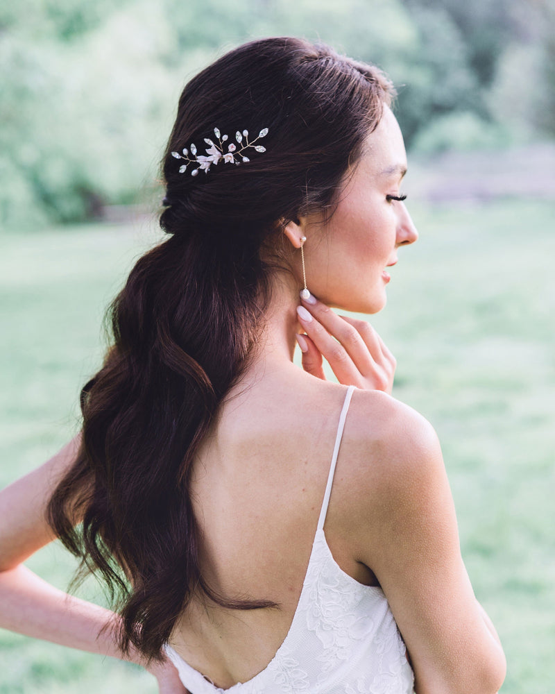 A side view of a dark-haired bride with long hair wearing a hair pin in the side of her half up hairstyle. She is also wearing long earrings with pearls at the bottom of a delicate chain.