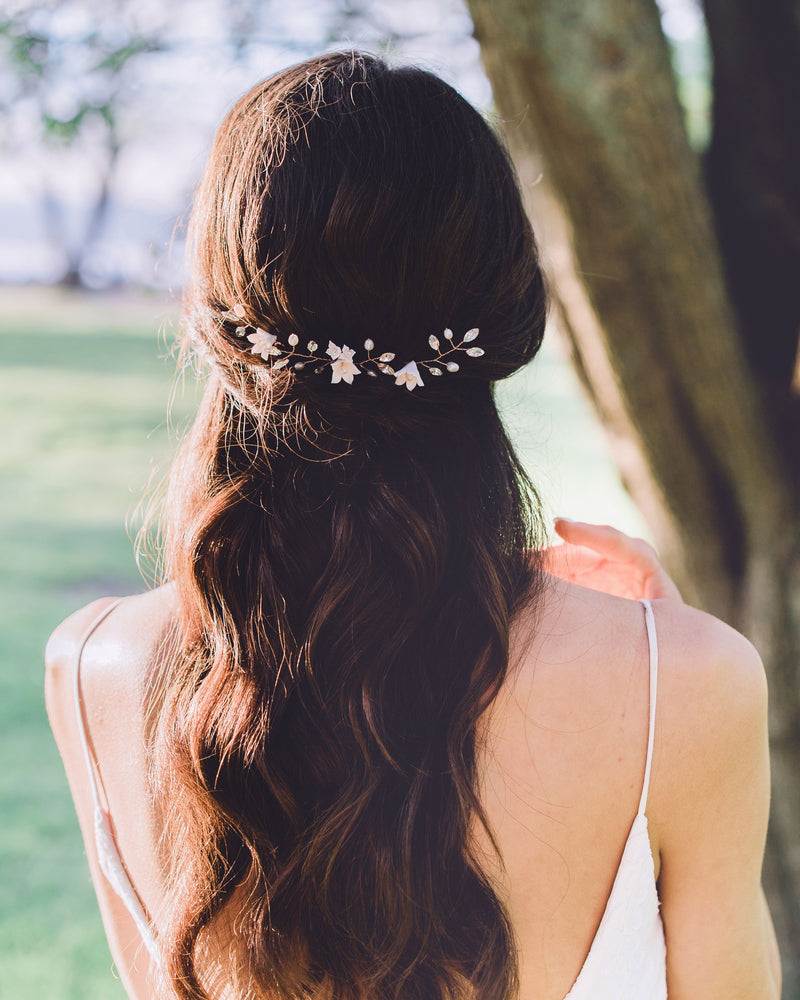A back view of a bride with long dark hair wearing a trio of hair pins in her half up hair. The hair pins have been placed along the back of her updo, so that it styles across the back of her hair. The hair pins are rose gold, with blush flowers, pearls, and crystals.