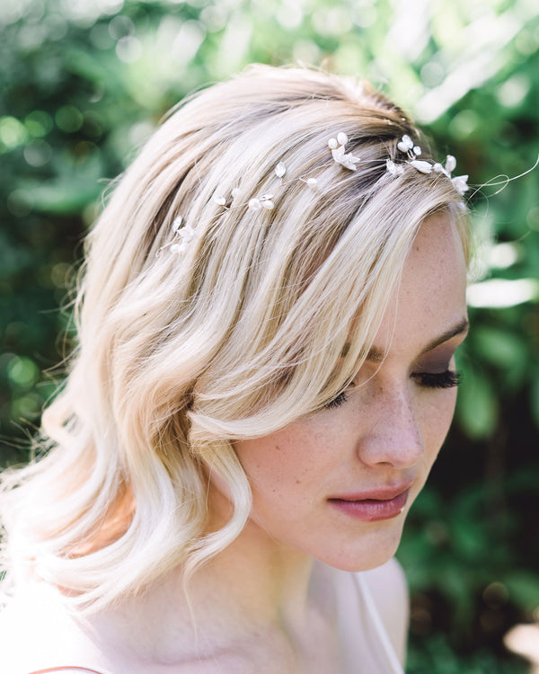 Model smiling softly and gazing downwards. She is wearing the delicate Belle Fleur bridal Hair Vine in gold made of dainty flowers, pearls, and crystals; styled to the front with soft waves in her hair.