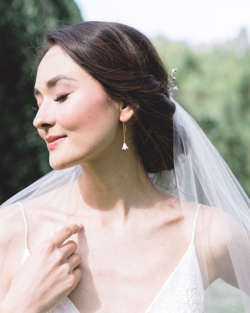 A dark-haired bride smiles with her eyes closed and turns her face to the sun. She is wearing delicate ear threader earrings with tiny blush flower drops. A veil is styled into her low updo.