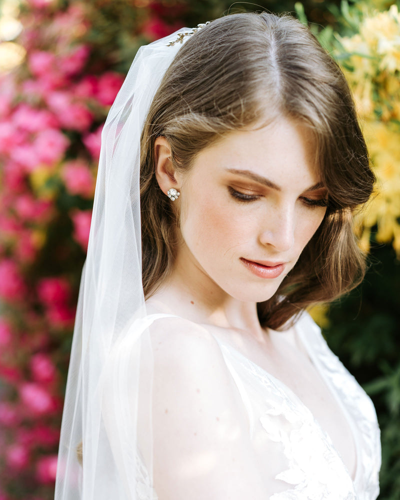 A model gazes down and shows a delicate crystal stud in her ear. She is also wearing a simple tulle veil in her auburn hair.