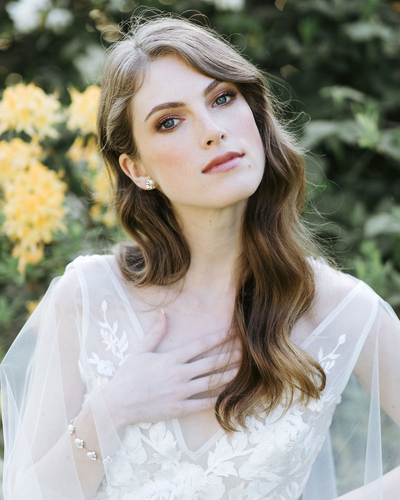 A bride wears a tulle bridal cape. She is modelling the Celestial Pearl Cluster Bracelet and Celestial Pearl Stud Earrings.