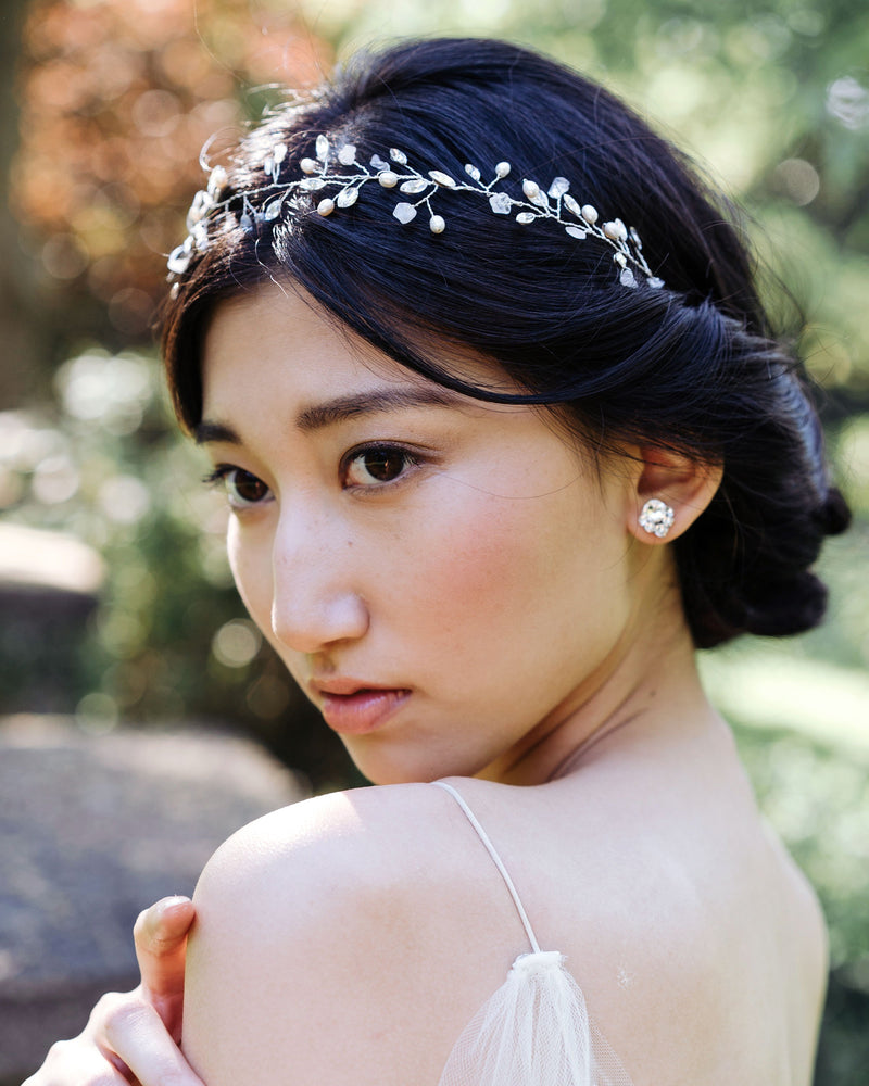 A bride gazes into the distance. She wears crystal stud earrings. Her black hair is styled in a low updo with twists and a silver hair vine with gemstones, pearls, and crystals.