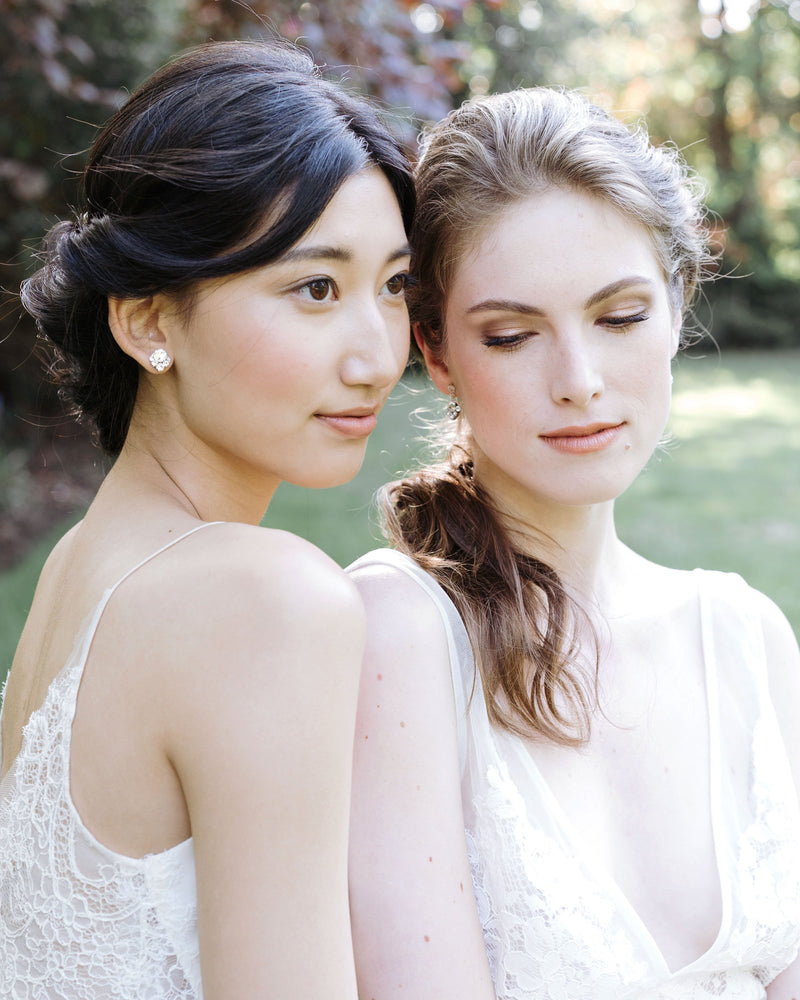 Two models wear bridal earrings. The dark-haired model on the left wears the Celestial Crystal Stud Earrings. The model on the right wears the drop version.