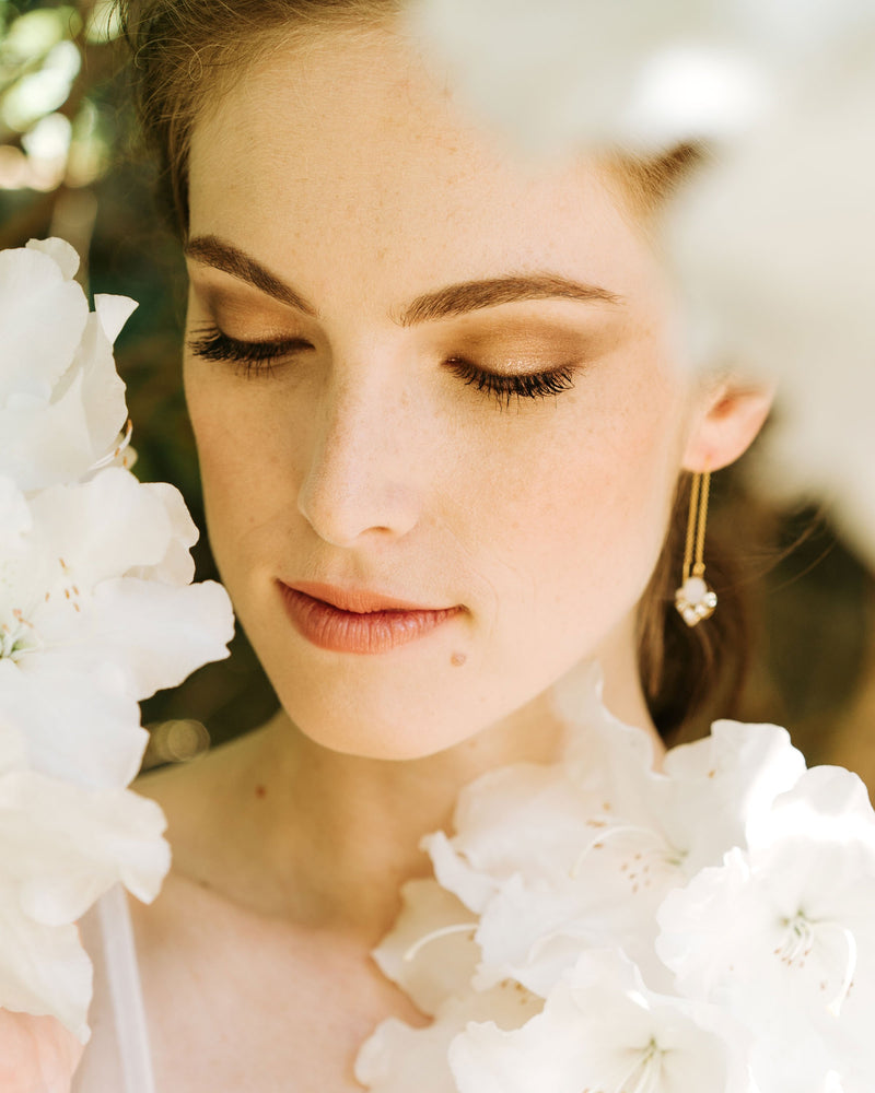 A model poses with white flowers all around her. She is wearing the Celestial Threader Earrings in gold with white opal crystals.