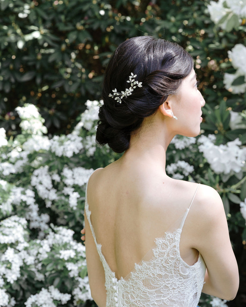 A dark-haired bride with a low bridal updo is wearing a crystal bridal comb made of crystal leaf clusters, styled to the side.