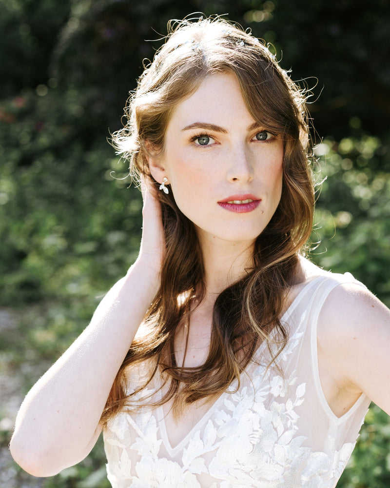 A bride with auburn hair stands with sun on her soft bridal waves. She is wearing the crystal leaf earrings in gold.