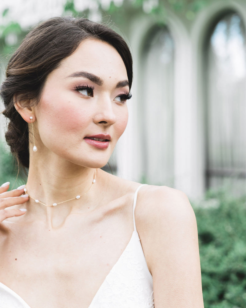 A model wears her dark hair in a low bridal updo. She is wearing the Dainty Pearl Necklace and the 2" Teardrop Pearl Long Earrings in gold.