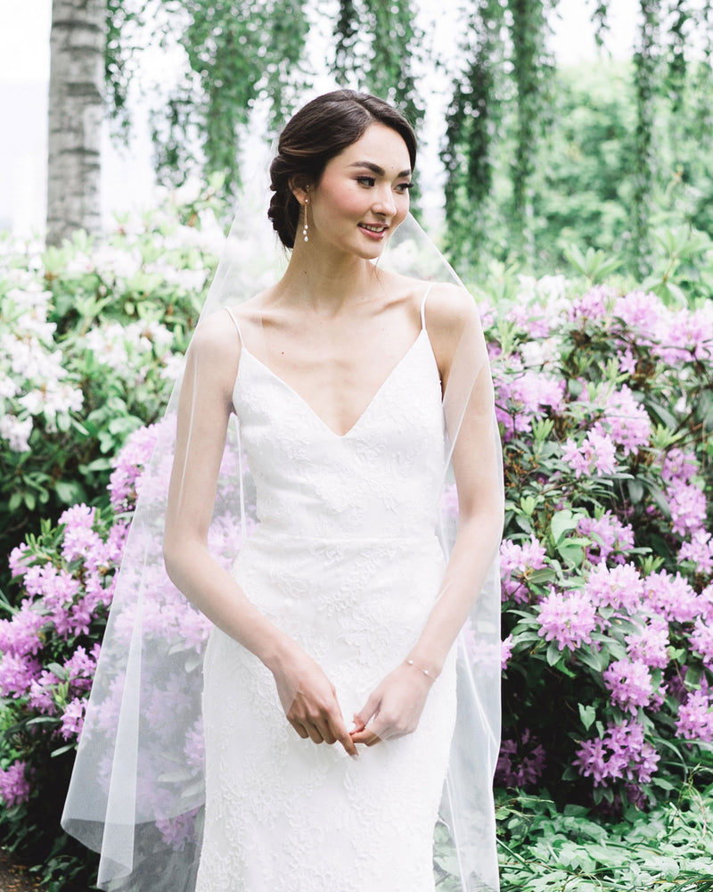A bride stands in front of blooming shrubs. She wears a soft veil without gathers, the Dainty Pearl Trio Earrings and matching Dainty Pearl Bracelet.