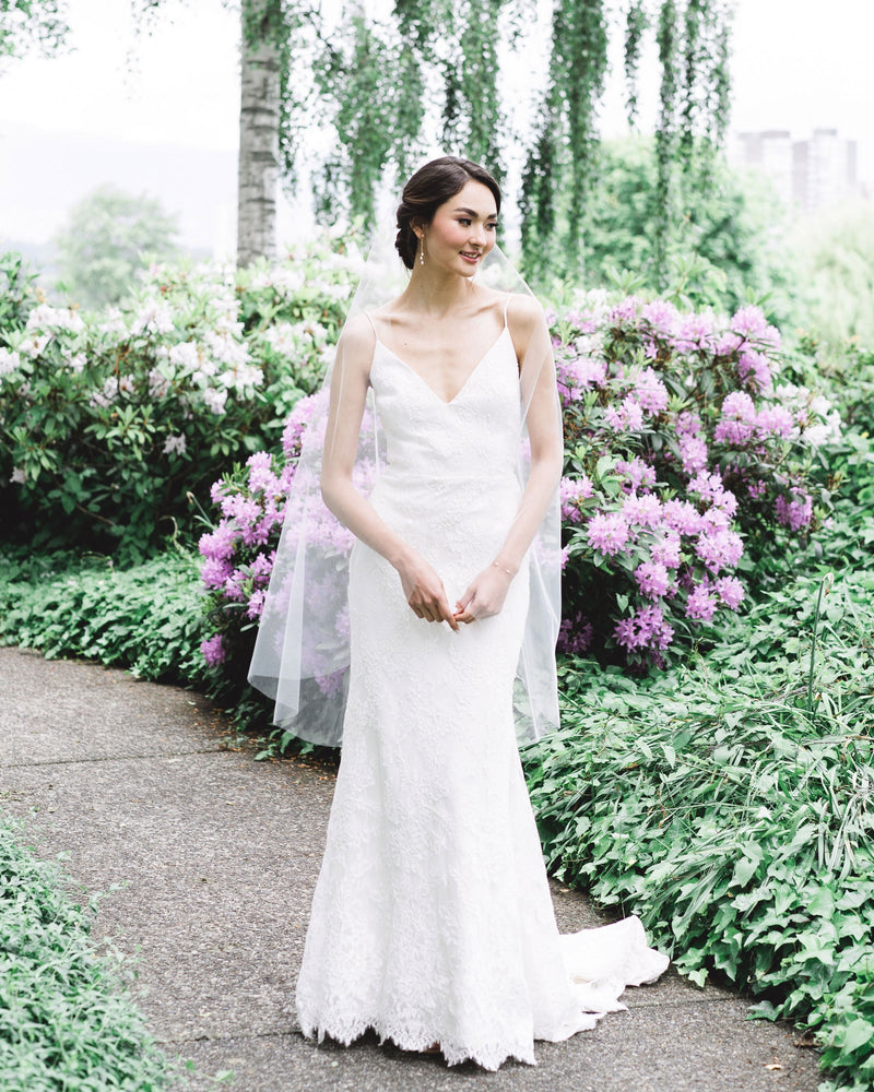 A bride stands in front of blooming shrubs. She wears a soft veil without gathers, the Dainty Pearl Trio Earrings and matching Dainty Pearl Bracelet.