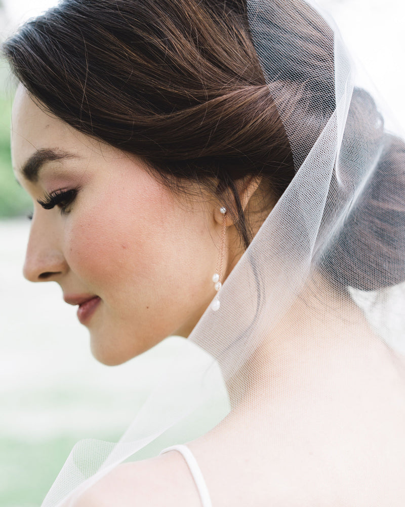 A bride wears delicate drop bridal earrings in rose gold, paired with a veil without gathers.