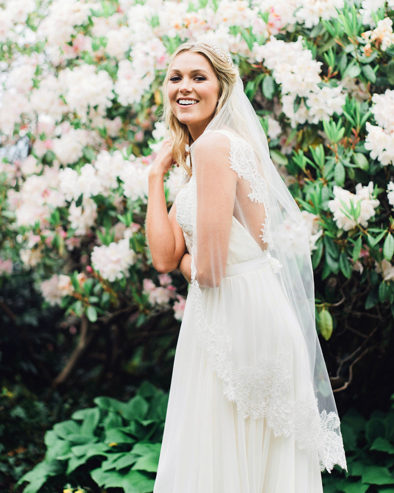 A blonde model smiles in a wedding dress. She is wearing a fingertip length chantilly lace veil.