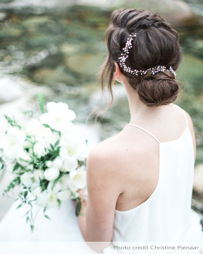 A bride sits along a rocky shore, with her hair styled in a braided low bun. A dainty hair vine with pearls and crystals is woven into her hair.