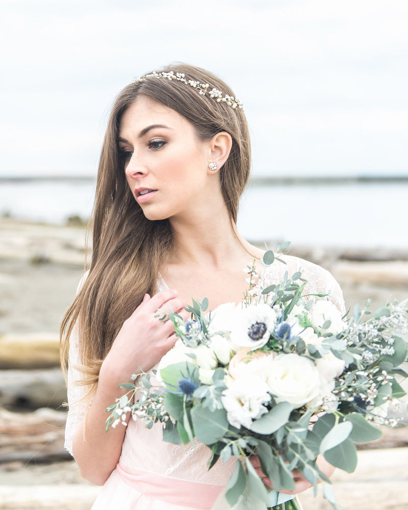 A bride holds a bouquet and wears a romantic golden halo of crystals and pearl clusters. Her long flowing locks are brushed over one shoulder.