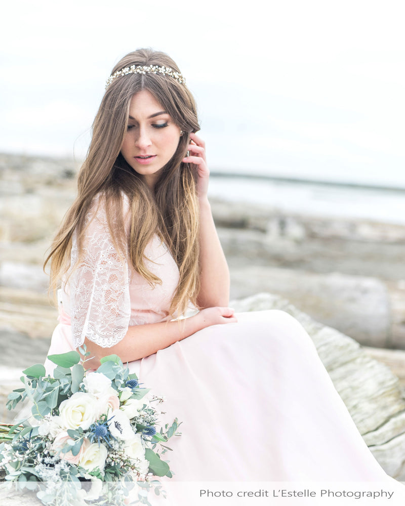 A bride poses at a rocky seashore. She wears a bridal halo of crystals and pearl clusters atop her long hair.