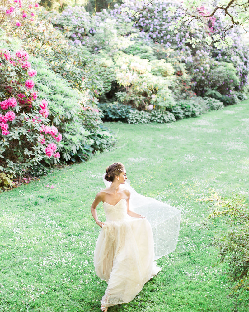 A bride runs across a grassy field with her veil floating softly behind her. She wears the Delphine two-layer veil in chapel length.