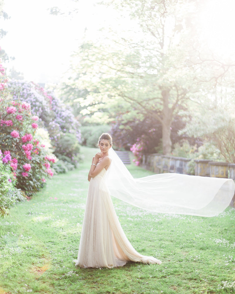 A model poses at golden hour with her veil floating softly behind her. She is wearing our Delphine two-layer veil in chapel length.