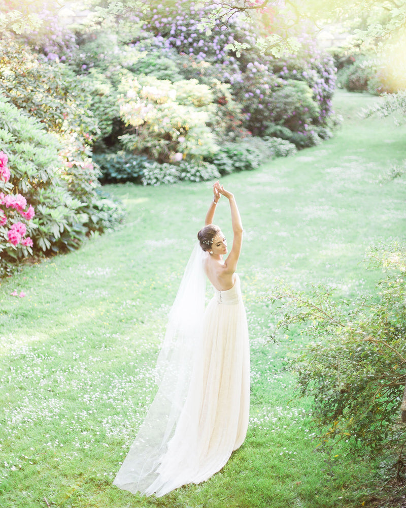 A bride dances in a bright, grassy field. Her soft two-layer veil trails behind her; she is wearing the Delphine Veil in chapel length.