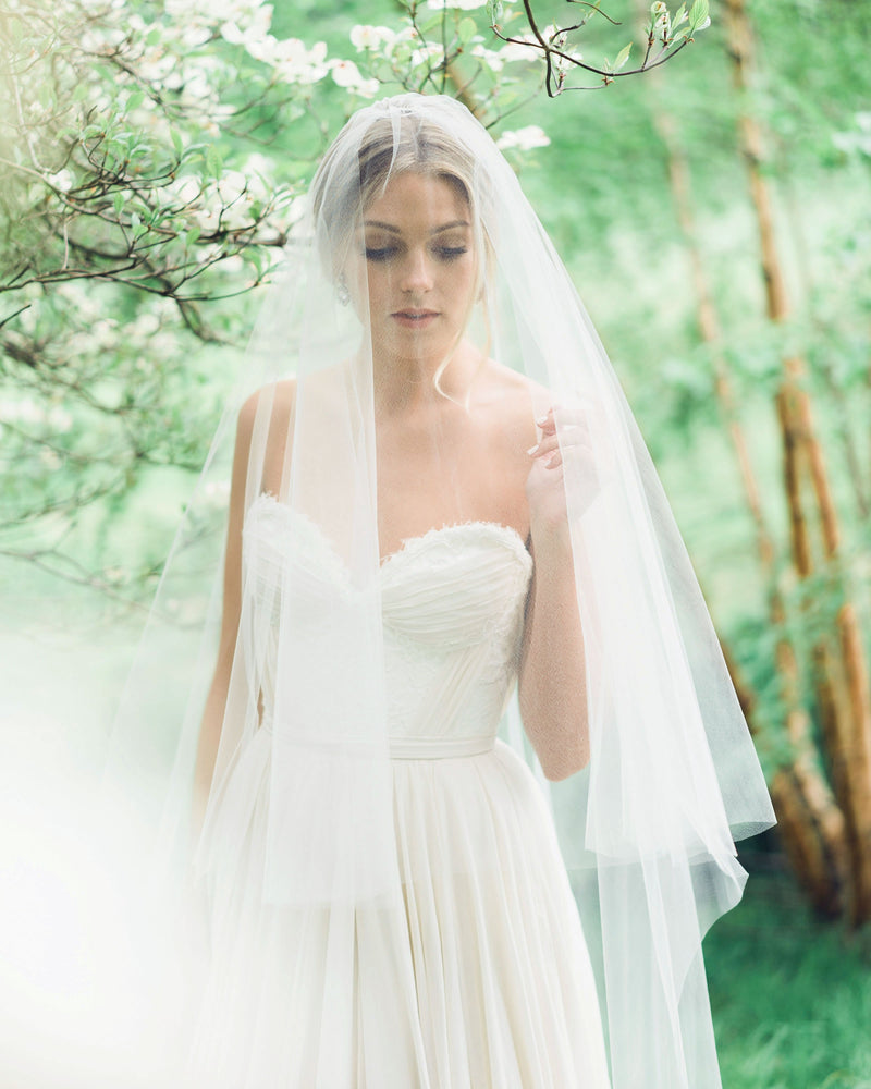 A bride poses in the forest. The blusher of her two-layer veil is worn to the front. She is wearing the Delphine Gathered Veil in fingertip length.