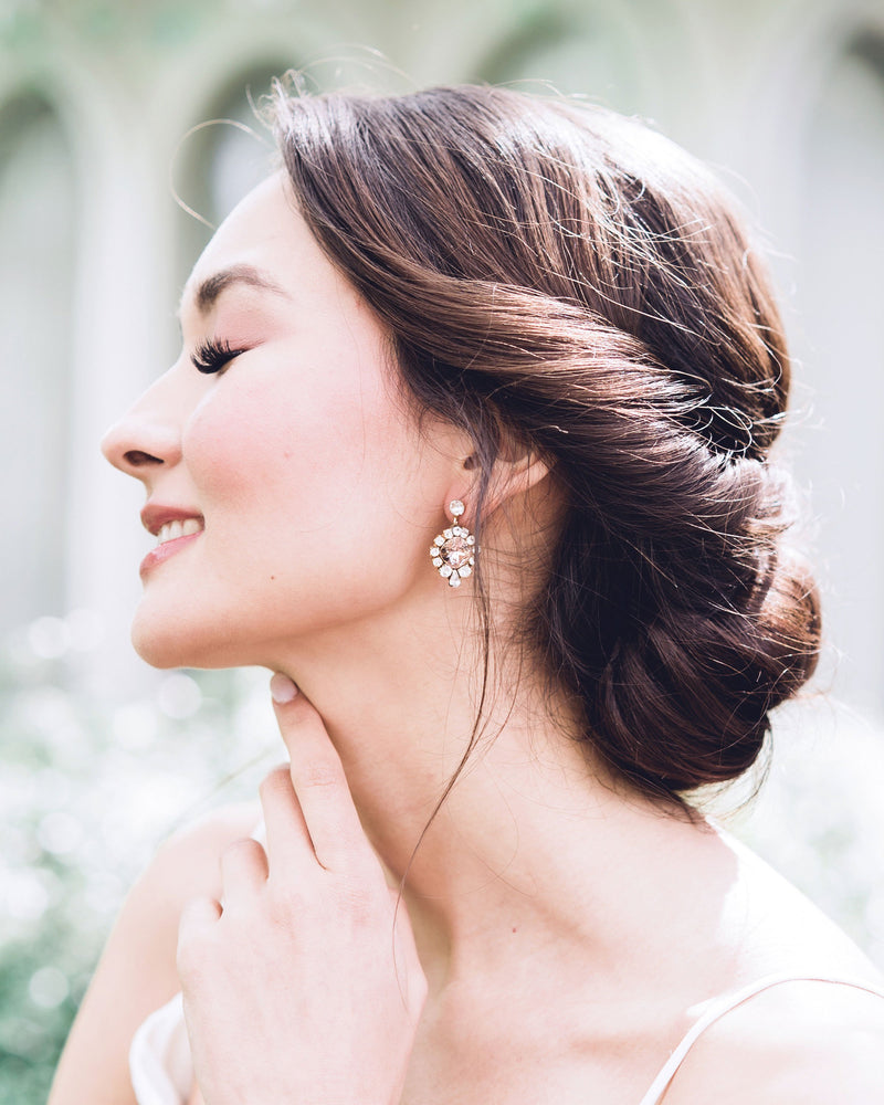 A model smiles as she wears the Enchanted Crystal Drop Earrings in gold with blush crystal centres. Styled with a low bridal updo with twists.