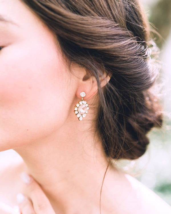 A close model view of a bride wearing the Enchanted Crystal Drop Earrings in gold with blush crystal centres. Styled with a low bridal updo with twists.