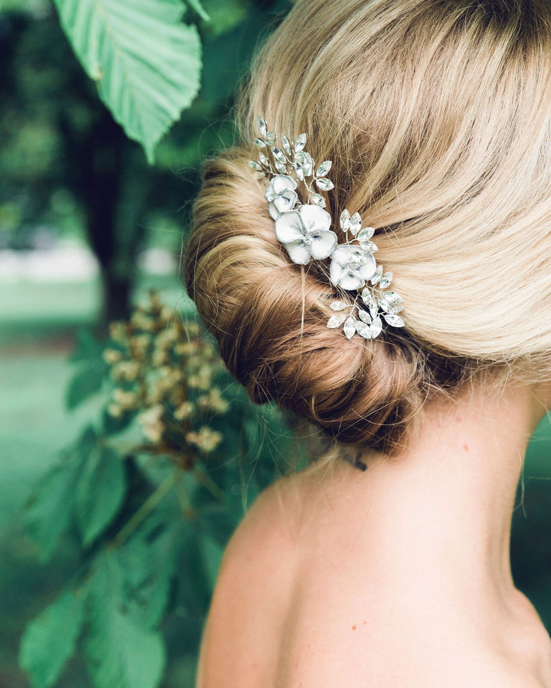 A blonde model has her hair styled into a modern bridal French twist. The Gilded Blossoms Comb is styled into the updo. The comb has white flowers with silver edges, pearls, and crystals.