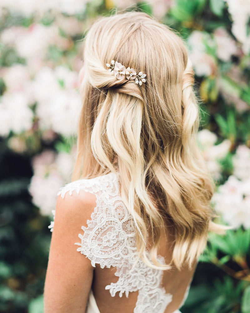 A bride has her hair styled in soft waves with both sides pinned back. She wears the Gilded Blossoms Petite Comb styled into the side. The comb has blush hand-painted flowers, pearls, and crystals.