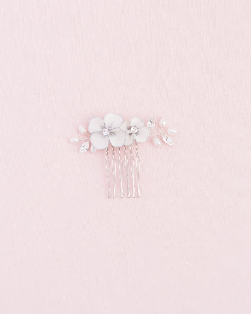 Flatlay on a pink background of the Gilded Blossoms Petite Comb in silver. The comb has white flowers hand-painted with a silver edge, pearls, and crystals.