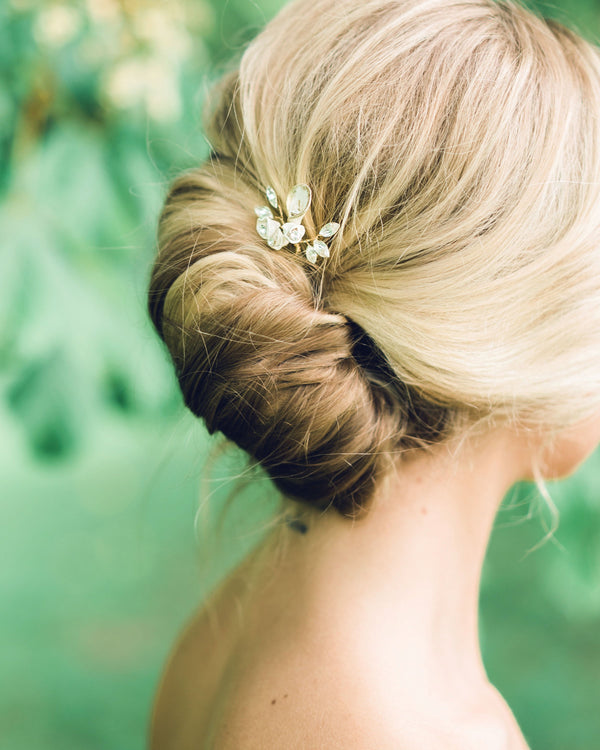 A blonde model has her hair styled into a modern bridal French twist. The Gilded Blossoms Comb is styled into the updo. The comb has white flowers with gold edges, pearls, and crystals.