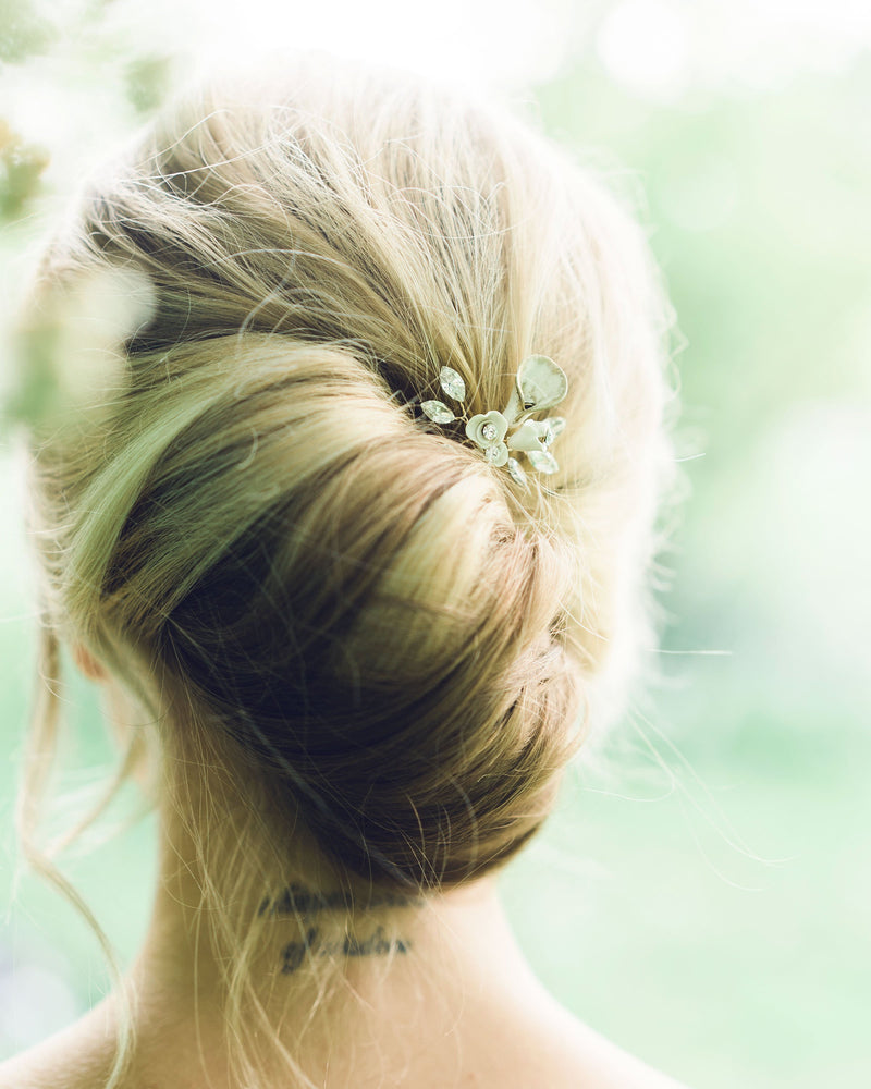 A blonde model has her hair styled into a modern bridal French twist. The Gilded Lily Hair Pin is styled into the updo. The hair pin has white flowers and crystals.